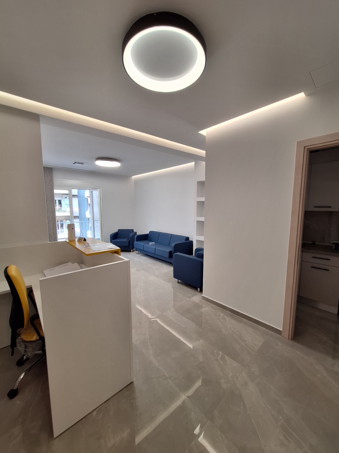 Modern office lounge with blue sofas, white reception desk, recessed lighting, and circular ceiling light.