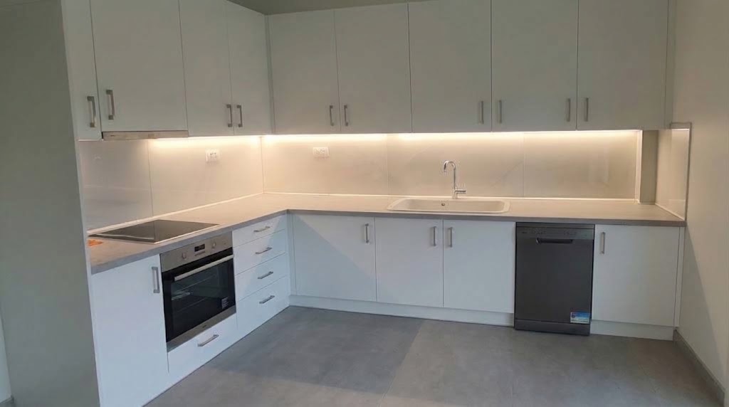 Modern L-shaped kitchen with white cabinets, built-in oven, sink, and dishwasher under bright lighting.